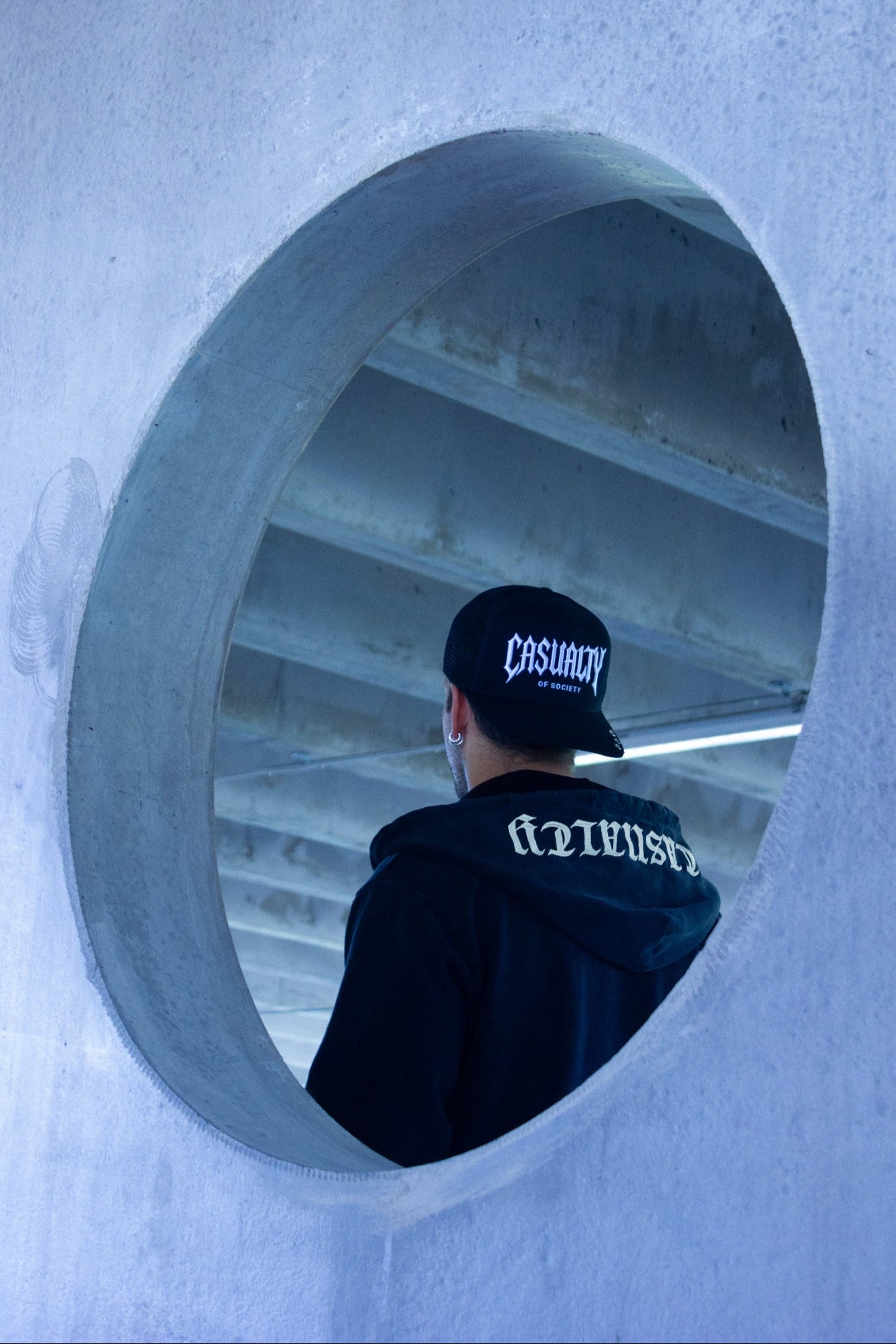 Person wearing a black cap and hoodie looking into a circular concrete opening.